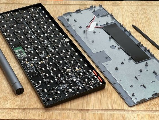 Opened mechanical keyboard with its internal circuitry exposed, lying next to its detached back cover on a wooden surface. A little green wireless PCB stands out from the black of the main PCB.