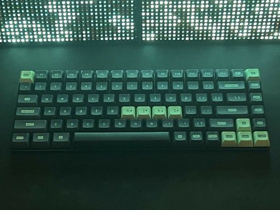 A mechanical keyboard with green and black keys.  The VIM h, j, k and l keys, arrow keys and Esc are picked out in neon green. A brightly lit LED matrix panel sits in the background with a game of life pattern displayed on it.