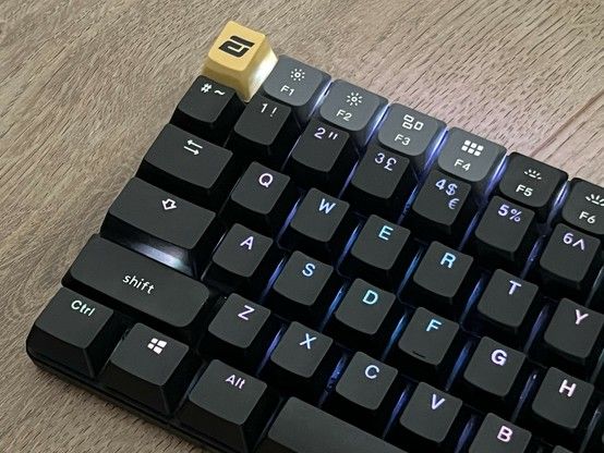 A close-up of a low profile mechanical keyboard with replacement full size black keycaps and customizable backlighting. The Escape key is highlighted with a yellow keycap featuring a custom symbol. The keycaps look… off… as if some of the non alphanumeric ones are placed in the wrong location. The F1 keys also have original, low profile caps on.