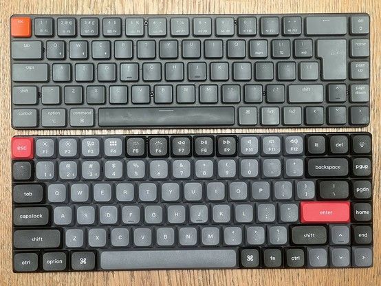 Two different mechanical keyboards on a wooden surface. The upper keyboard is dark grey with a reddish orange 