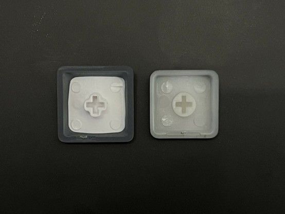 Two mechanical keyboard keycaps shown upside down against a dark background. The left one is slightly larger and looks less well made, with a a sort of blobby misshapen rectangle of white for the stem and legend.