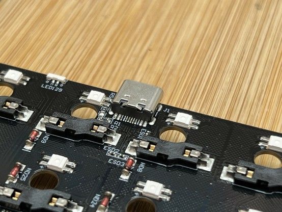 A close-up of a black printed circuit board with switch components, small white LEDs, and a USB-C connector, set against a wooden background.
