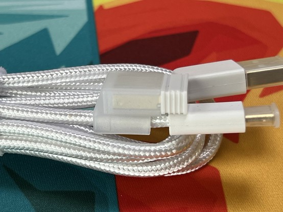 Close-up of a white braided cable with USB connectors on a colorful background. A small plastic clip attaches a separate USB transmitter to the cable for storage, in an effort to prevent it getting lost.