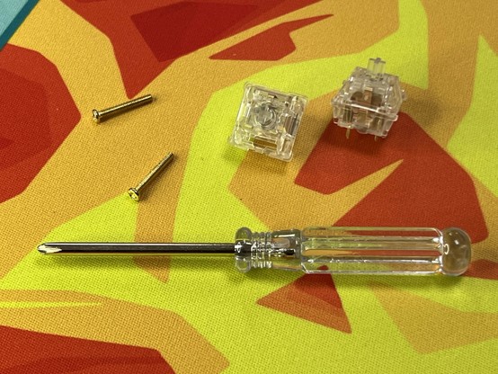 A clear plastic screwdriver, two golden screws, and two clear plastic mechanical keyboard switches on a colorful red, orange and yellow background.