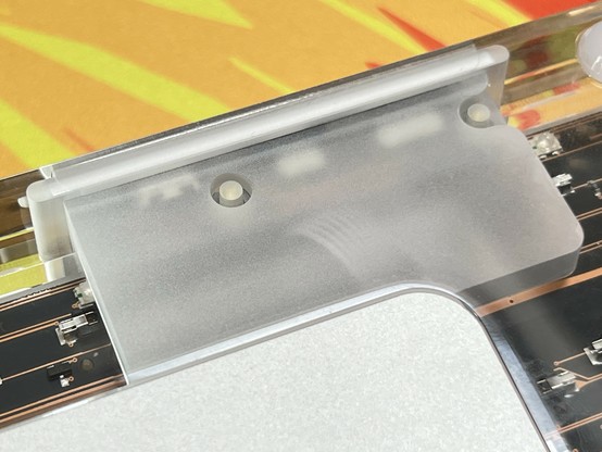 Close-up of a frosted section of the transparent acrylic case on a mechanical keyboard. The backs of some switches and ports, plus a ribbon cable can be faintly seen through it. Set against a colorful, blurred, red, orange and yellow background.