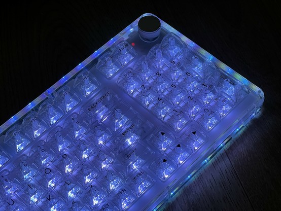 A transparent mechanical keyboard with blue LED backlighting on a dark surface. Around the edge is a green and blue striped effect from the LEDs underneath.
