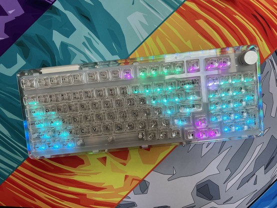 A transparent mechanical keyboard with RGB backlighting placed on a colorful, abstract-patterned desk mat.