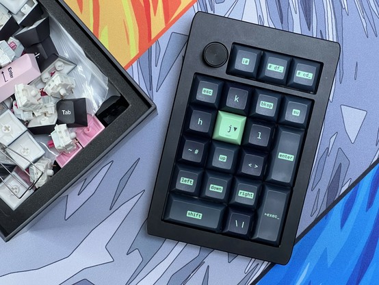 A box of loose keyboard switches, caps and associated tools next to a custom keypad with black and green keys on a colorful desk mat.