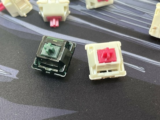 Close-up of several mechanical keyboard switches, with emphasis on a green switch and a reddish pink/white switch.