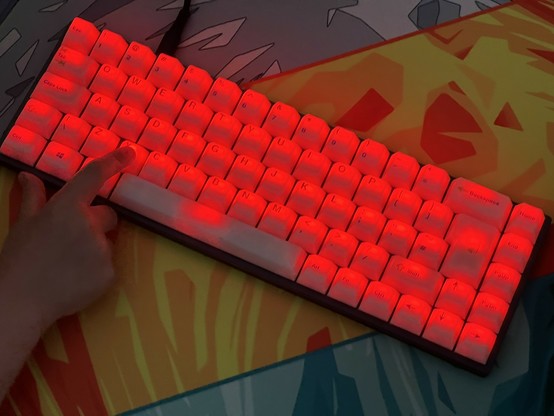 A close-up of a mechanical keyboard with red backlighting, placed on a colorful desk mat. A finger is pressing a key. The key caps are completely transluscent with printed text so they’re all lighting up like red jellies.