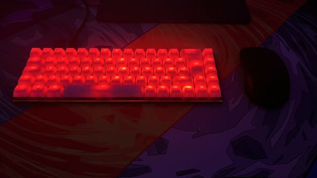 A mechanical keyboard with fully transluscent illuminated red keys and a black mouse on a colorful desk mat. The keycaps have bright spots and non uniform lighting, and the less said about the sad one LED below the spacebar the better… but it looks okay…
