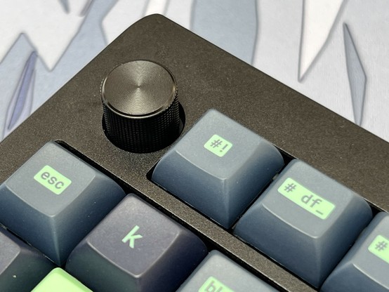 Close-up of a mechanical keyboard with deep blue keycaps and a black volume knob. The focus is on a #! Key which used to read i# 🫠