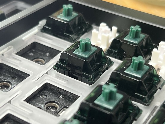 Close-up image of green mechanical keyboard switches pushed into a plastic plate. The focus is on a switch that’s just slightly proud of the plate though it’s not obvious.