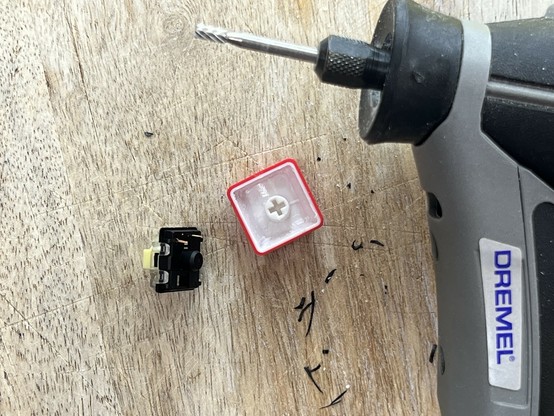 A Dremel tool is shown with a drill bit attached, next to a keyboard switch and a keycap on a wooden surface.