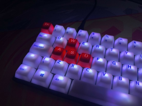 A mechanical keyboard with illuminated keys, featuring red keycaps on the WASD keys and the Escape key. It’s lit up with white light which makes the configuration very evocative of a certain national flag…