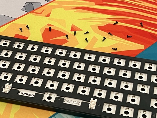 A disassembled mechanical keyboard on a colorful mat. Thirteen screws are messily set aside.