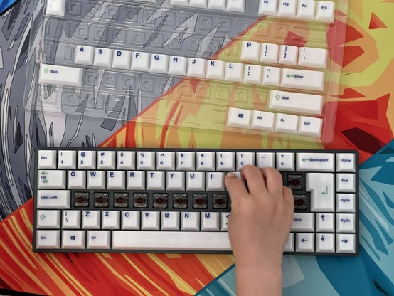 A child's hand is placing keycaps on a mechanical keyboard with some keycaps missing. A keycap set and a colorful abstract mat are visible in the background.