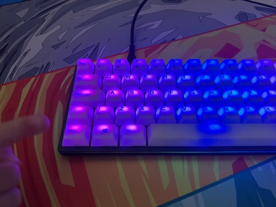 A mechanical keyboard with purple and blue backlighting on a colorful desk mat. A little hand is pointing to the left side of the keyboard.
