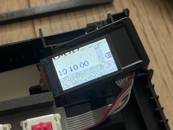 Close-up of mechanical keyboard with a small LCD screen displaying distorted numbers, a cut off battery icon and a confetti static background.