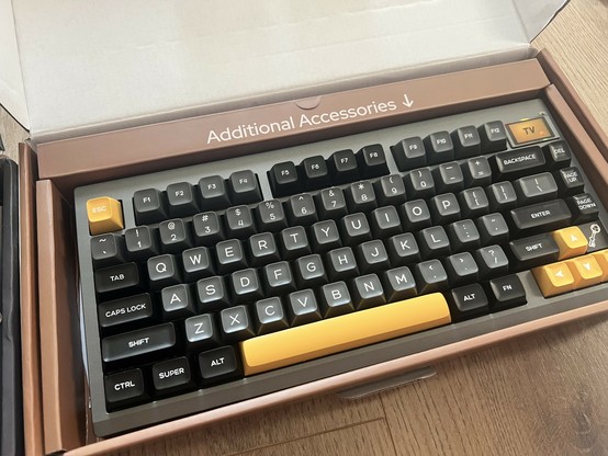 A mechanical keyboard with black, grey and yellow keys in a tan brown box. The keys are scooped like a 1980s home computer keyboard.