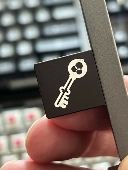 Close-up of fingers holding a black plastic thingy with a white vintage key symbol. There’s a blurred keyboard in the background.