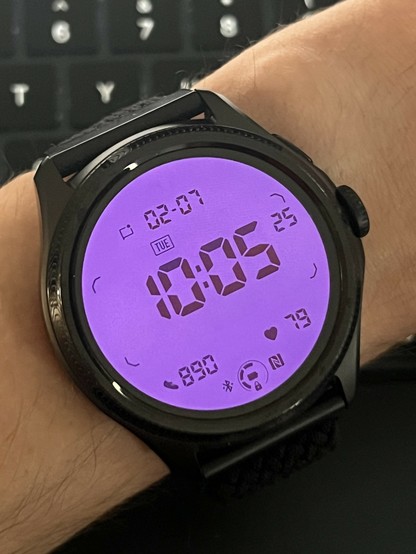 A smartwatch showing the date and time with a low power low fidelity seven segment styled TN display. It’s backlit pink with the OLED behind it.