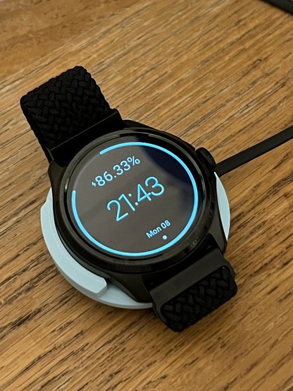 A smartwatch with a black strap is charging on a wooden surface. The screen displays the battery percentage at 86.33%, the time as 21:43, and the date as 