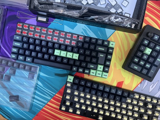 Custom mechanical keyboard parts, tools, and kits on a colorful desk mat. There are two distinct but near identical keyboards with keycaps being moved from one to the other.