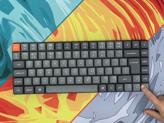 A mechanical keyboard with grey keys, an orange 'esc' key, and a multicolored desk mat underneath. A finger is pressing the 'end' key.