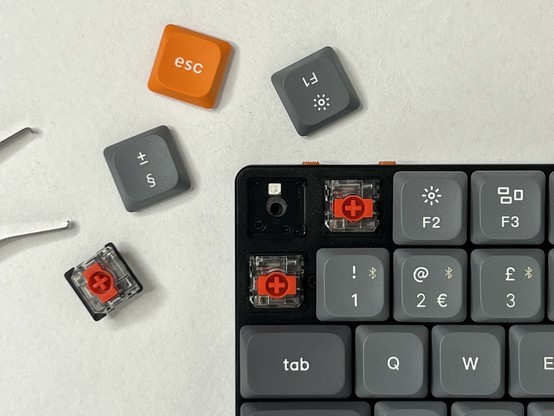 Close-up of a mechanical keyboard with several keycaps removed, revealing red switches beneath. Loose keycaps, a removed switch and a switch puller are placed beside the keyboard.