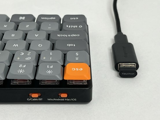 A close-up of a mechanical keyboard with grey and orange keycaps, displaying part of the key layout. Alongside is a USB cable with a USB C to A adapter and a dongle inserted labeled 