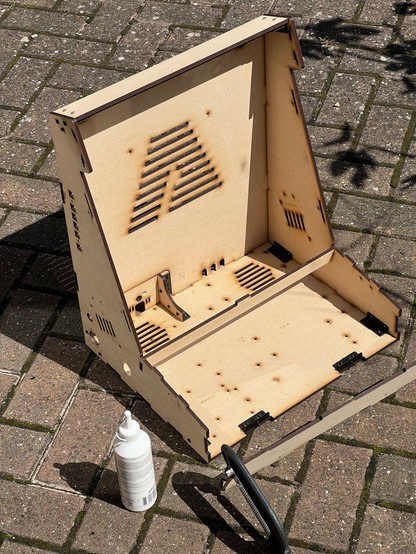 Partially assembled MDF arcade cabinet case on a paved surface, accompanied by a bottle of wood glue and a clamp.