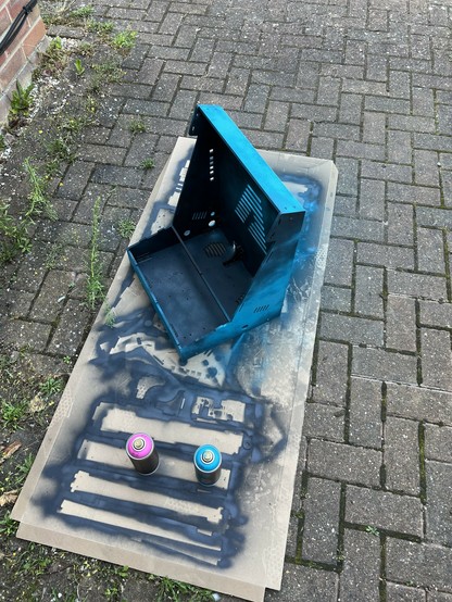 A wooden bartop arcade cabinet chassis partly spray-painted blue on a piece of cardboard on a brick pavement. Two spray paint cans, one pink and one blue, are also on the cardboard.