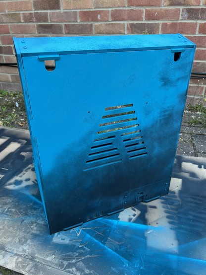 An arcade cabinet painted black with a rough layer of blue shading up to solid blue near the top, placed on a surface with splattered paint and a brick wall in the background.