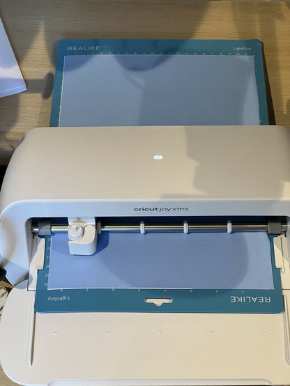 A Cricut Joy Xtra cutting machine with a blue REALIKE LightGrip mat on a wooden surface. On the mat is stencil fill being cut.