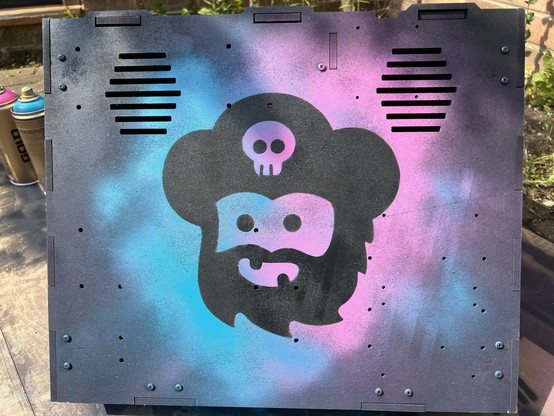 Spray-painted stencil art on a black surface, featuring a stylized skull with a hat and beard. The background is a mix of purple and blue spray paint.