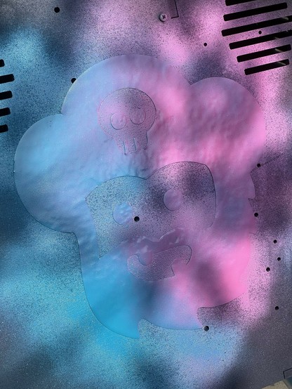 Spray paint covering a stencil of a stylized, cartoon-like face with a skull symbol on its head,  using blue and pink colors on a textured background.