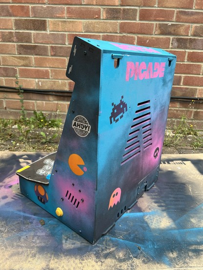 Custom arcade cabinet with retro gaming theme featuring neon colors, real stencilled graffiti artwork, and joystick controls, set against a brick wall background. A Pac-Man and Mario mushroom are visible on the left hand side. A pac-man ghost and space invader on the back.