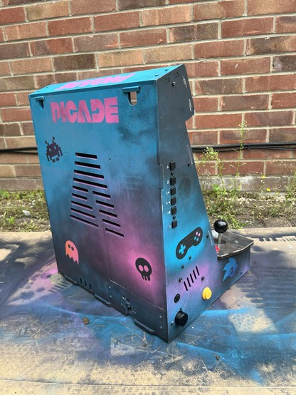 Custom arcade cabinet with retro gaming theme featuring neon colors, real stencilled graffiti artwork, and joystick controls, set against a brick wall background. A sonic logo and a gamepad icon are visible on the right-hand side, with silver paint pen labels for the LCD controls.