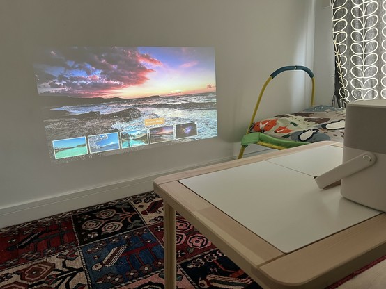 Image of a wall projection displaying a sunset beach scene barely a foot above the floor. The source is a small white projector on a wooden kids play table.