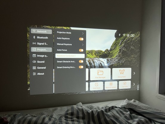 A projector screen displaying various settings and menu options such as Network, Bluetooth, Projection Mode, Auto Keystone, and more. There is a backdrop of a waterfall scene, and the projection is on a wall above a bed with a rumpled duvet.