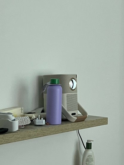 A shelf with a projector, a purple bottle, a lotion dispenser, an electrical plug and other detritus.