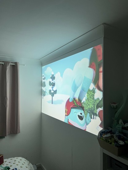 A projected image of an animated blue character in a red hat on a wall in a room. The room has light-colored curtains, a bed with white sheets, and a shelf with an embarrassment of unsorted odd kids socks.