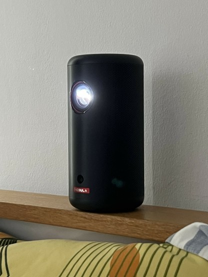 Small black cylindrical projector on a wooden bed headboard with a bright light coming from the lens.