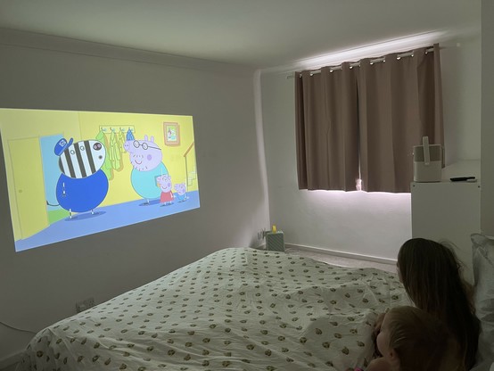 A bedroom with a projector screen displaying an animated cartoon featuring animal characters. Two children are sitting on a bed watching the cartoon. The room has light-colored walls and curtains. The projector is stood on a high unit next to the bed.