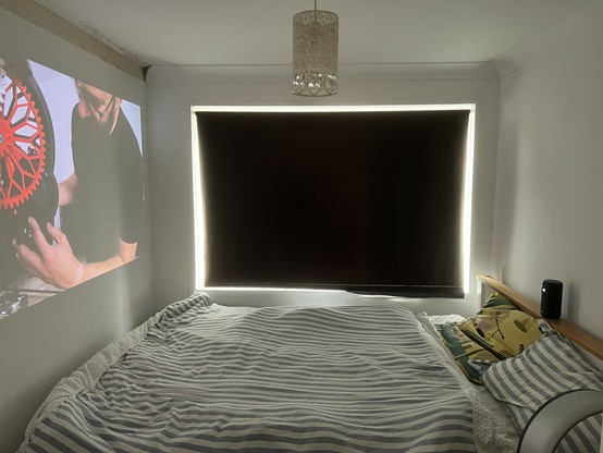 A bedroom with a striped bedspread, a window with a dark curtain, and a tiny projector throwing a screen displaying a video of a person working on a mechanical device.