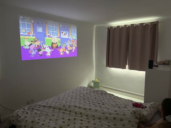 A dimly lit bedroom with a projector screen displaying an animated children's show. A bed with patterned sheets is in the foreground, and a person is lying on the bed near a partially visible stuffed animal. Closed curtains cover the window. The projector is a tiny cylinder perched on a unit to the right.