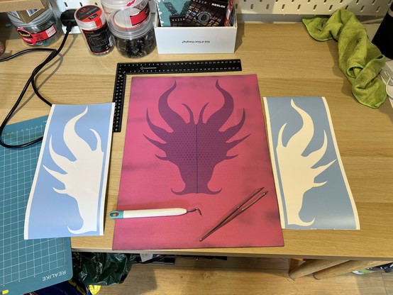 A dragon face stencil in two halves masking over a pink base coat on a canvas. The stencil film pieces with the shapes removed sit either side of the canvas. A weeding tool and pointy tweezers sit on top. A ruler sits above for dialling in the alignment.