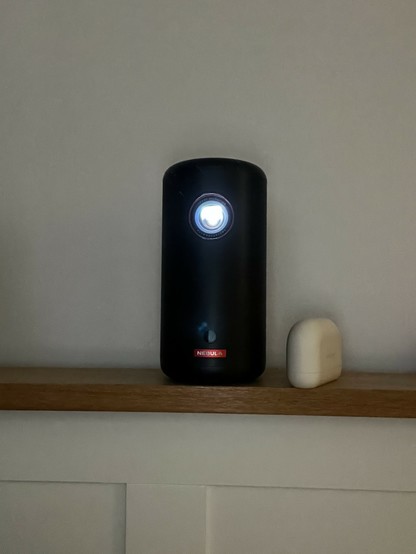 An image of a black Nebula projector with a bright light at the top, placed on a wooden shelf next to a white Jabra Elite 10 earbuds case, against a light-colored wall. The projector appears to be about 3-4x as tall as the earbuds case.