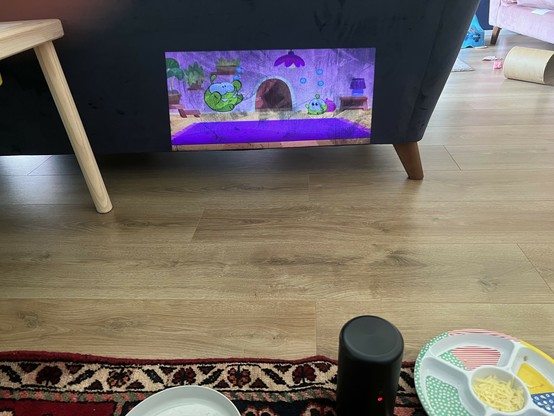 A cozy room scene featuring a projector displaying an animated image of cute green characters projected on the back of a dark blue couch. The projector sits in the foreground, along with a divided plate with food items. The floor is wooden, and a patterned rug is visible beneath the plate. The projected image is so clear it looks photoshopped on.